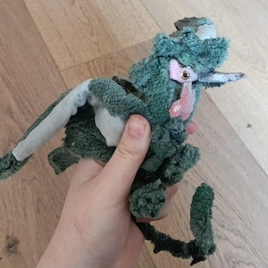 Peut inclure: Une peluche faite à la main, dans les tons de vert et de bleu-vert, avec un accent rose. Le jouet a une longue queue et une forme de bec. La peluche est tenue dans une main, mettant en valeur sa taille et sa texture. La peluche semble être une créature fantaisiste.