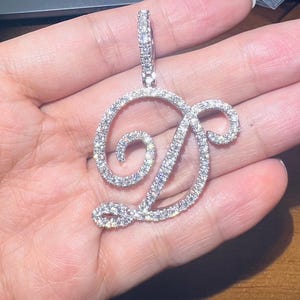 May include: A sparkling, silver-toned pendant in the shape of the letter "D", encrusted with numerous small, clear gemstones. The pendant has a loop at the top for a chain. The design is elegant and suitable for jewelry.