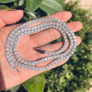 May include: A sparkling, silver-toned necklace with numerous clear, round-cut gemstones. The necklace is held in a hand, with a clasp visible. The background features green foliage, suggesting an outdoor setting. The gemstones reflect light, creating a brilliant display.