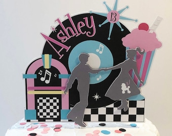 May include: A white cake with a pink, blue, and black 50s themed cake topper. The topper features a record with the name "Ashley" on it, a jukebox, a dancing couple, and a cupcake. The cake is decorated with pink, blue, and black confetti.