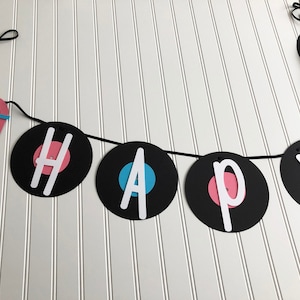 May include: A banner with the word "HAPPY" spelled out on black vinyl record cutouts. The letters are white, with pink and blue accents. A jukebox-shaped tag is attached to the banner.