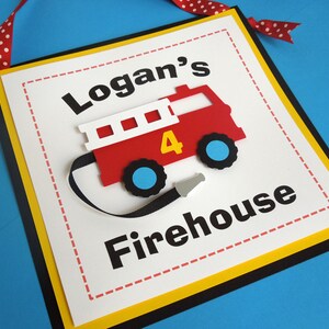 May include: A white sign with a yellow border and black trim. The sign has a red fire truck with a black hose and the words "Logan's Firehouse" in black.