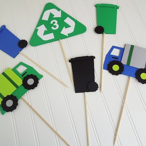 May include: A set of six green, blue, and black recycling themed cupcake toppers. The toppers include a recycling sign with the number 3, a blue recycling bin, a green recycling bin, a black recycling bin, and two green recycling trucks.