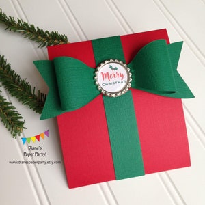 May include: A red paper gift box with a green ribbon and bow. The bow is centered on the box and has a decorative silver bottle cap with the words "Merry Christmas" printed on it. The background is white.