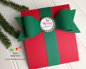 May include: A red paper gift box with a green ribbon and bow. The bow is centered on the box and has a decorative silver bottle cap with the words "Merry Christmas" printed on it. The background is white.