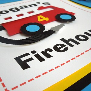 May include: A white sign with red dashed lines around the border. The sign says "Logan's Firehouse" in black letters. A red paper fire truck with black wheels and a black ribbon is attached to the sign.