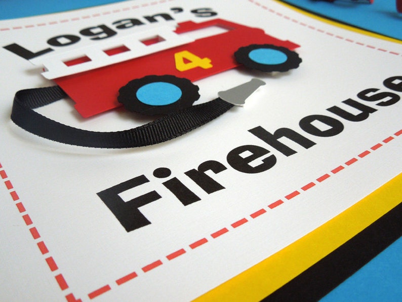 May include: A white sign with red dashed lines around the border. The sign says "Logan's Firehouse" in black letters. A red paper fire truck with black wheels and a black ribbon is attached to the sign.