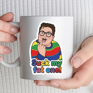 Könnte beinhalten: Weiße Keramiktasse mit einem Aufkleber mit einer Cartoon-Illustration einer Person mit Brille und einem gestreiften Pullover, die den Mittelfinger zeigt. Der Text "Suck my fat one!" befindet sich darunter.