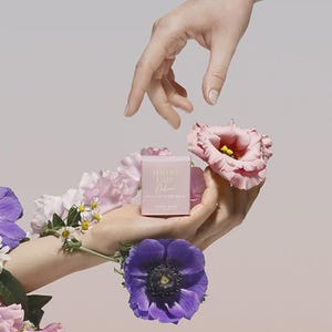 May include: A pink box of Tender Care Multi-Purpose Balm, held in a hand surrounded by pink and purple flowers. The text on the box is in a script font. The background is a soft, neutral colour.