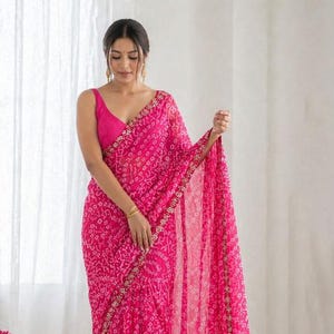 May include: A vibrant pink saree with a delicate pattern and gold trim. The saree is draped over a matching pink blouse. The fabric appears lightweight and flowing, with intricate detailing along the borders.
