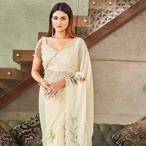 May include: A cream-coloured saree with a floral design. The saree has a scalloped edge and a painted floral pattern in pink, green, and blue. The woman is wearing a cream-coloured top with a V-neck.