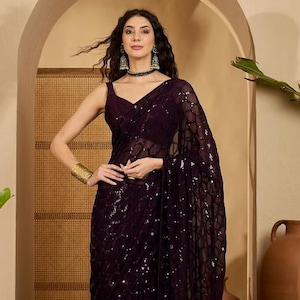 以下が含まれることがあります： スパンコール模様のダークパープルのサリー。ノースリーブのブラウスとシアーで流れるような生地が特徴です。モデルは、カフブレスレットやイヤリングなど、ゴールドのジュエリーを身に着けています。背景には、装飾的なアーチと模様のあるラグがあります。