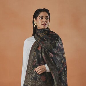 May include: A black shawl with a floral pattern and a fringed edge is draped over a person wearing a white long-sleeved top and skirt. The shawl features a detailed design with red, orange, and green accents. The background is a solid orange color.