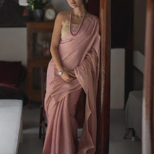 Könnte beinhalten: Eine Frau trägt einen hellrosa Sari und eine passende Halskette. Der Sari ist mit weichen Falten und dezentem Glanz drapiert. Sie steht in der Nähe einer Holzsäule und trägt eine Uhr. Der Hintergrund zeigt einen Raum mit einem Gemälde und Pflanzen.