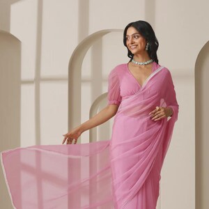 May include: A light pink saree with a sheer, flowing fabric. The saree features a V-neckline and short sleeves. The edges are trimmed with a delicate white border. The model is wearing a matching necklace and earrings.