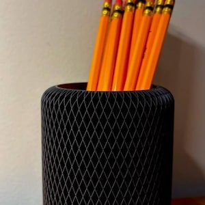 May include: A black, cylindrical pencil holder with a diamond-patterned design, filled with bright orange pencils. The pencils have pink erasers and gold-colored metal bands. The holder sits on a wooden surface.
