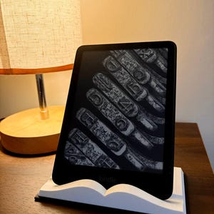 May include: A black Kindle e-reader displaying a close-up of typewriter keys, resting on a white book-shaped stand. The stand is on a dark wooden surface, with a lamp in the background.