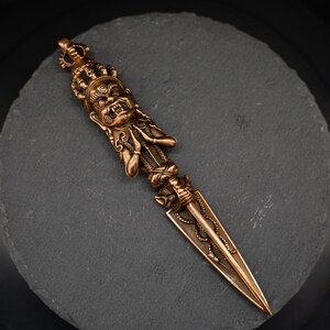 May include: Bronze-colored ritual dagger with intricate details. The handle features a detailed face and ornate carvings. The blade is triangular with a serpent design. The dagger is displayed on a dark gray, circular surface.