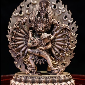 Op de afbeelding: Een bronzen metalen standbeeld dat een godheid met meerdere armen voorstelt in een dynamische omhelzing met een kleinere figuur. De centrale figuur heeft meerdere hoofden, een woeste uitdrukking en talloze armen die zich uitstrekken tegen een grote, versierde, vlamvormige halo.
