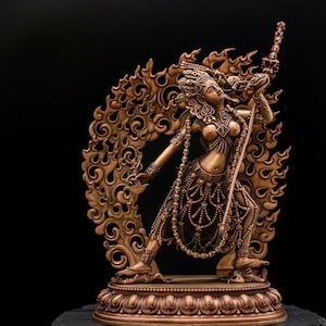 Op de afbeelding: Bronskleurig beeld van een vrouwelijke figuur met sierlijke details, die een staf vasthoudt. Het beeld heeft een decoratieve achtergrond en staat tegen een zwarte achtergrond. De figuur is versierd met sieraden en ingewikkelde patronen.