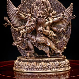 May include: A bronze-coloured statue depicting a multi-armed deity in a dynamic pose, embracing a consort. The figure features multiple heads, wings, and intricate ornamentation, standing on a lotus-petal base with small figures beneath the feet.
