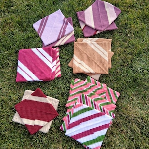 May include: An overhead shot displays a collection of colorful, square, knitted coasters. The coasters feature various stripe patterns in shades of pink, purple, beige, and green. The coasters are arranged on a bed of green grass.