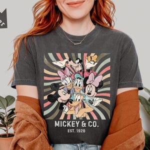 May include: Dark gray t-shirt featuring a colorful graphic of Mickey Mouse, Minnie Mouse, Donald Duck, Daisy Duck, Goofy, and Pluto. The shirt has the text "Mickey & Co. Est. 1928" in a vintage style.