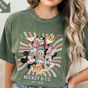 Puede incluir: Camiseta verde oliva con un dise&ntilde;o de Mickey Mouse, Minnie Mouse, Donald Duck, Daisy Duck, Goofy y Pluto. El dise&ntilde;o est&aacute; sobre un fondo a rayas con el texto "Mickey & Co. EST. 1928".