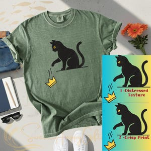 May include: Green t-shirt with a black cat graphic dropping a gold crown. The shirt is styled with blue jeans, a notebook, sunglasses, and white sneakers. A graphic shows the cat design with a distressed texture and a crisp print.