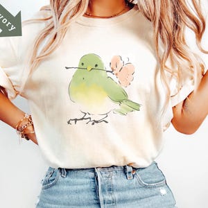 May include: Ivory-colored t-shirt featuring a watercolor illustration of a bird holding a twig and pink flowers. The bird is green and yellow. The shirt is paired with blue denim shorts. The word "Ivory" is printed on a green arrow.