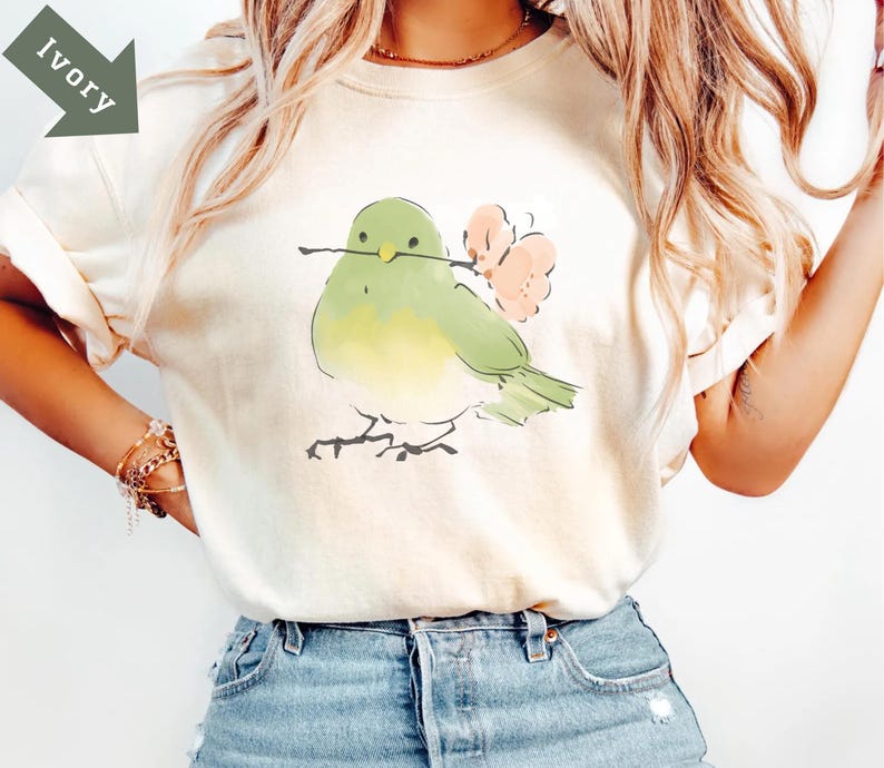 May include: Ivory-colored t-shirt featuring a watercolor illustration of a bird holding a twig and pink flowers. The bird is green and yellow. The shirt is paired with blue denim shorts. The word "Ivory" is printed on a green arrow.
