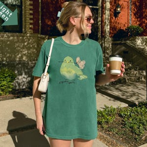 May include: A light green t-shirt featuring a watercolor illustration of a bird with a twig in its beak and two pink butterflies. The shirt is a relaxed fit and has a round neckline. The model is wearing a white purse and holding a coffee cup.