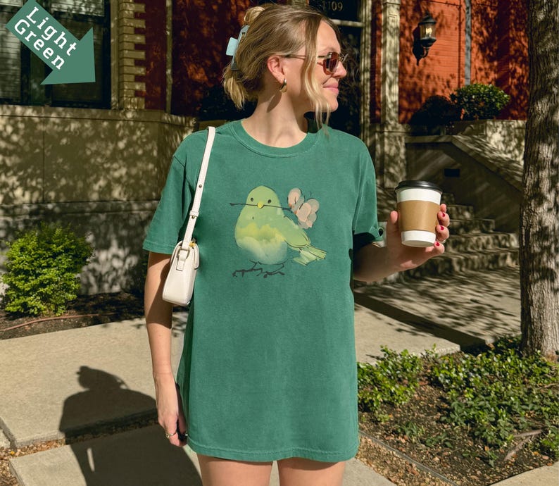 May include: A light green t-shirt featuring a watercolor illustration of a bird with a twig in its beak and two pink butterflies. The shirt is a relaxed fit and has a round neckline. The model is wearing a white purse and holding a coffee cup.