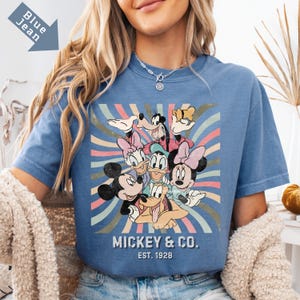 Puede incluir: Camiseta azul vaquero con un gr&aacute;fico de Mickey Mouse y sus amigos. El dise&ntilde;o incluye a Mickey, Minnie, Donald, Daisy, Goofy y Pluto, con un fondo retro. El texto "MICKEY & CO. EST. 1928" est&aacute; impreso debajo de los personajes.
