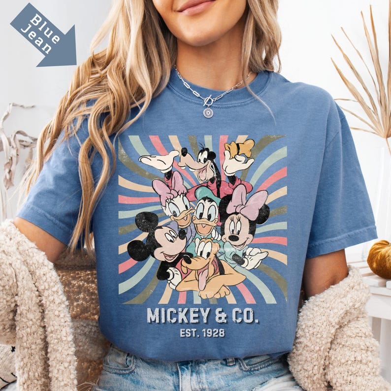 Puede incluir: Camiseta azul vaquero con un gr&aacute;fico de Mickey Mouse y sus amigos. El dise&ntilde;o incluye a Mickey, Minnie, Donald, Daisy, Goofy y Pluto, con un fondo retro. El texto "MICKEY & CO. EST. 1928" est&aacute; impreso debajo de los personajes.