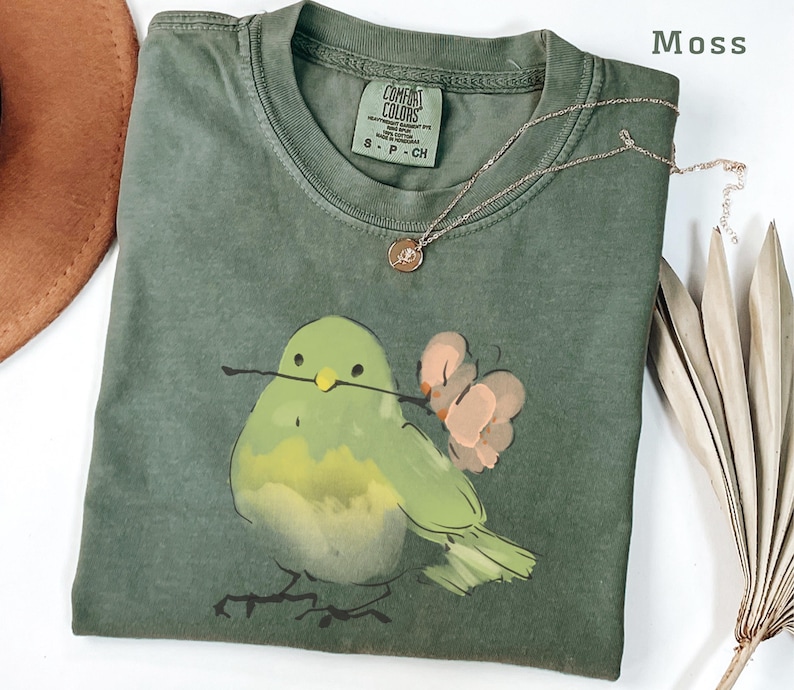 May include: A moss green t-shirt with a round neck and short sleeves. The shirt features a watercolor illustration of a green bird holding a twig and pink flowers. A gold necklace with a pendant is draped on the shirt.