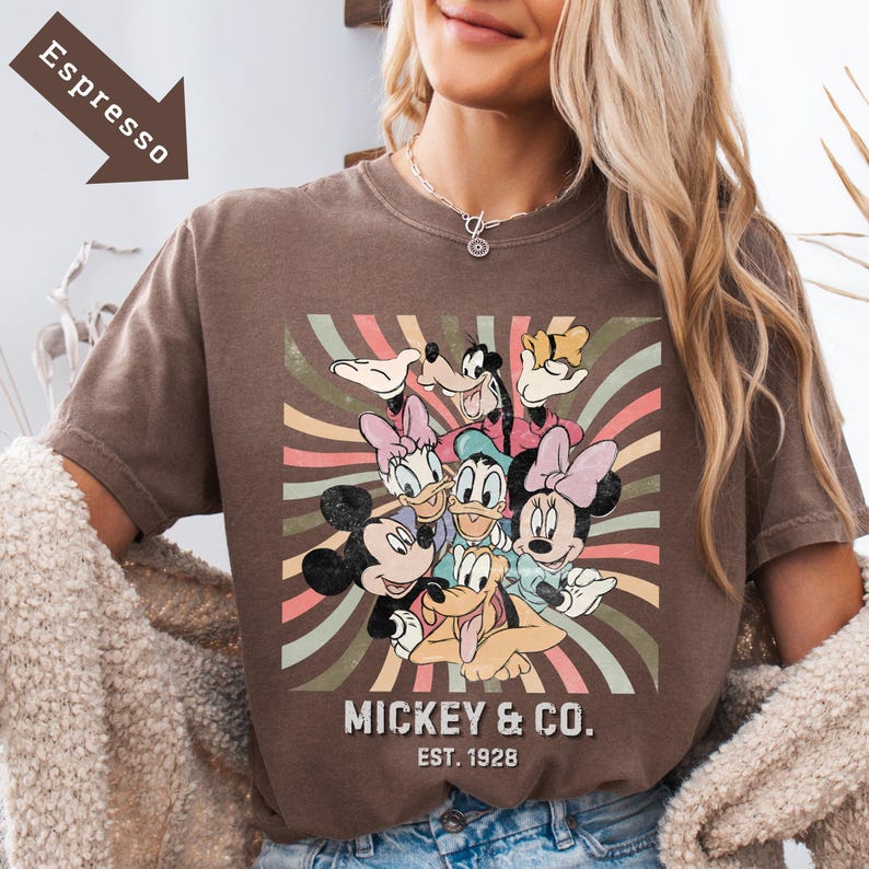 Puede incluir: Camiseta color espresso con un gr&aacute;fico de Mickey Mouse y sus amigos. El dise&ntilde;o incluye a Mickey, Minnie, Donald, Daisy, Goofy y Pluto, con el texto "Mickey & Co. Est. 1928". El fondo tiene un dise&ntilde;o de rayas retro.