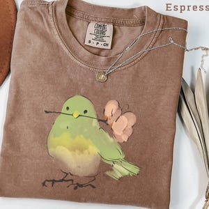 May include: Espresso-colored t-shirt with a watercolor illustration of a green bird holding a twig and flowers. The shirt has a crew neck and a small tag. A silver necklace with a pendant is draped across the shirt.