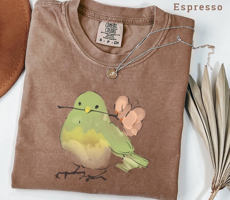 May include: Espresso-colored t-shirt with a watercolor illustration of a green bird holding a twig and flowers. The shirt has a crew neck and a small tag. A silver necklace with a pendant is draped across the shirt.