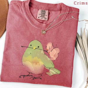 May include: Crimson-colored t-shirt with a watercolor illustration of a bird holding a twig and flowers. The shirt has a crew neck and a small tag. A silver necklace with a pendant is draped across the shirt.