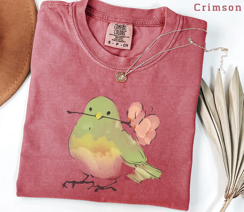 May include: Crimson-colored t-shirt with a watercolor illustration of a bird holding a twig and flowers. The shirt has a crew neck and a small tag. A silver necklace with a pendant is draped across the shirt.