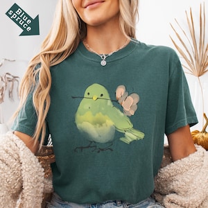 May include: A teal-colored t-shirt featuring a watercolor illustration of a green bird holding a twig and a pink flower. The text "Blue Spruce" is printed on a teal arrow in the upper left corner. The shirt has a crew neck.