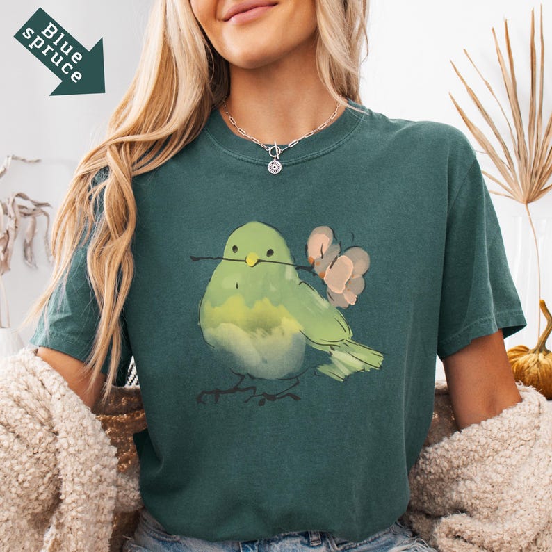 May include: A teal-colored t-shirt featuring a watercolor illustration of a green bird holding a twig and a pink flower. The text "Blue Spruce" is printed on a teal arrow in the upper left corner. The shirt has a crew neck.