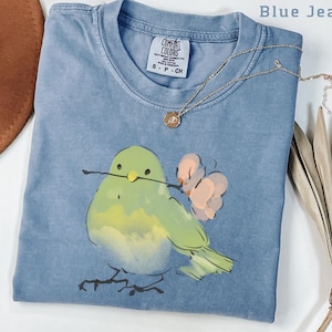 May include: Blue jean colored t-shirt with a watercolor illustration of a green bird holding a twig and a pink flower. A gold necklace with a pendant is on the shirt. The shirt has a Comfort Colors tag.