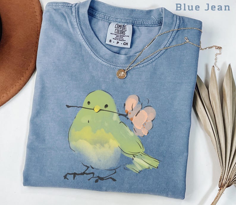 May include: Blue jean colored t-shirt with a watercolor illustration of a green bird holding a twig and a pink flower. A gold necklace with a pendant is on the shirt. The shirt has a Comfort Colors tag.
