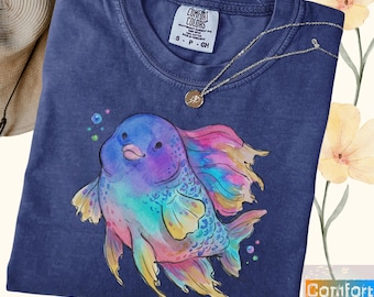 May include: A navy blue crew neck t-shirt featuring a watercolor-style illustration of a colorful fish with iridescent purple, blue, and pink scales and flowing fins. The shirt has a Comfort Colors label at the neck and a thin gold-tone necklace.