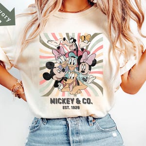 Puede incluir: Camiseta color marfil con un dise&ntilde;o retro de Mickey Mouse, Minnie Mouse, Donald Duck, Daisy Duck, Goofy y Pluto. El dise&ntilde;o est&aacute; sobre un fondo a rayas con el texto "Mickey & Co. Est. 1928".