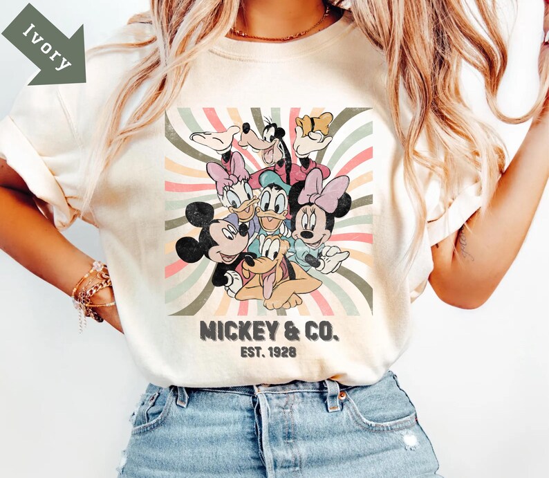 Puede incluir: Camiseta color marfil con un dise&ntilde;o retro de Mickey Mouse, Minnie Mouse, Donald Duck, Daisy Duck, Goofy y Pluto. El dise&ntilde;o est&aacute; sobre un fondo a rayas con el texto "Mickey & Co. Est. 1928".