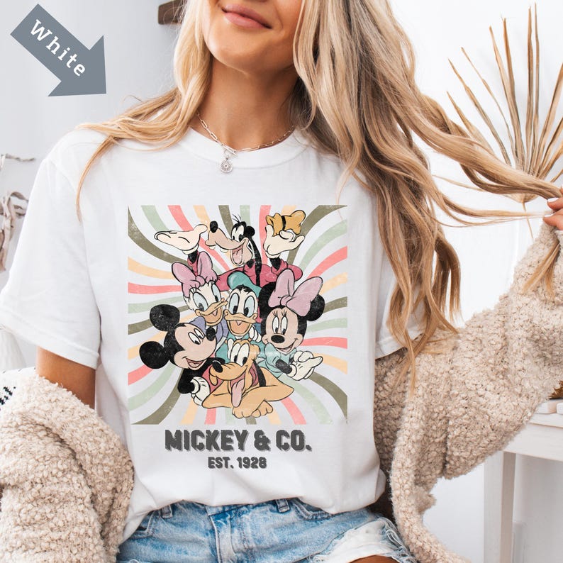 Puede incluir: Camiseta blanca con un gr&aacute;fico colorido de Mickey Mouse, Minnie Mouse, Donald Duck, Daisy Duck, Goofy y Pluto. El dise&ntilde;o incluye el texto "Mickey & Co. Est. 1928" en negro. El fondo tiene un dise&ntilde;o de rayas retro.