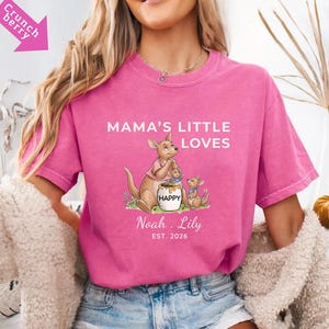 Puede incluir: Una camiseta rosa con el texto "MAMA'S LITTLE LOVES" y un dise&ntilde;o de familia de canguros de dibujos animados. El dise&ntilde;o incluye un canguro, dos cr&iacute;as y un tarro de miel con la palabra "HAPPY". Los nombres "Noah. Lily" y el a&ntilde;o "EST. 2026" tambi&eacute;n est&aacute;n impresos en la camiseta.
